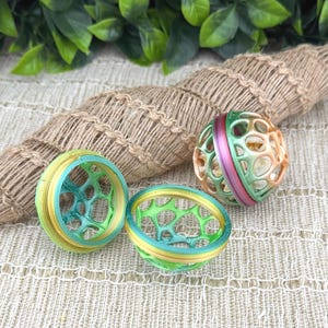 May include: Three colorful, spherical and ring-shaped objects. The spheres are green, orange, and pink. The rings are yellow, blue, and green. The objects have a geometric, open design. The background includes a woven rope and green leaves.