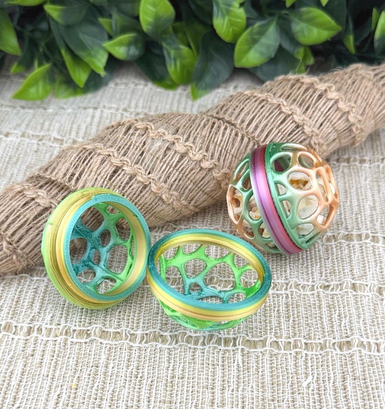 May include: Three colorful, spherical and ring-shaped objects. The spheres are green, orange, and pink. The rings are yellow, blue, and green. The objects have a geometric, open design. The background includes a woven rope and green leaves.