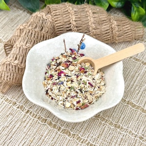 May include: A white, petal-shaped bowl filled with a mixture of dried herbs, flowers, and grains. A small wooden spoon rests in the bowl. The ingredients include pink rose petals and green herbs. The bowl sits on a tan, textured surface.