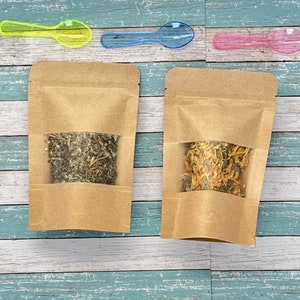 May include: Two brown paper pouches with clear windows, each filled with dried herbs. One contains green herbs, the other orange and yellow herbs. Three small plastic spoons in yellow, blue, and pink are above the pouches.