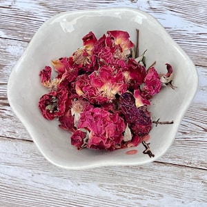 May include: A white ceramic bowl filled with dried pink rose petals. The petals are arranged in a loose pile and have a slightly wrinkled texture.
