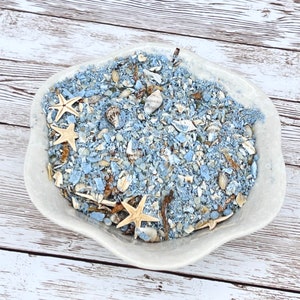 May include: A white ceramic bowl filled with blue and white sand, seashells, and starfish. The bowl is on a white wooden surface.