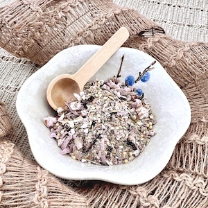 May include: A white, scalloped-edge bowl filled with a mix of grains, seeds, and dried fruits, accompanied by a small wooden spoon. The bowl is garnished with a sprig of blue berries. The bowl is set on a woven, textured surface.