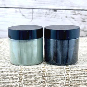 May include: Two clear jars with black lids, one filled with a light green powder and the other with a dark gray powder. The jars are sitting on a beige woven surface.