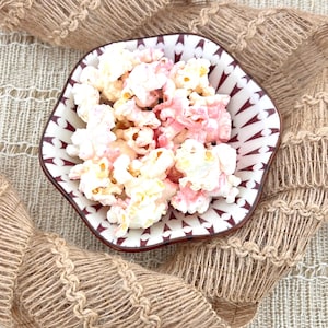 May include: A decorative, hexagonal bowl filled with popcorn. The popcorn is a mix of white and pink kernels. The bowl has a white interior with a brown and white geometric pattern around the rim. The background is a tan, woven fabric.