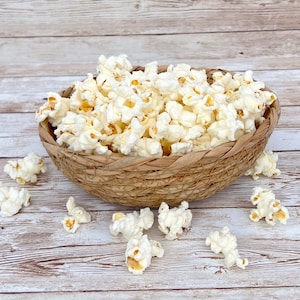 May include: A woven basket filled with fluffy, white popcorn. Several pieces of popcorn are scattered around the basket on a light-colored, wood-grain surface. The popcorn has a light yellow hue in the center.