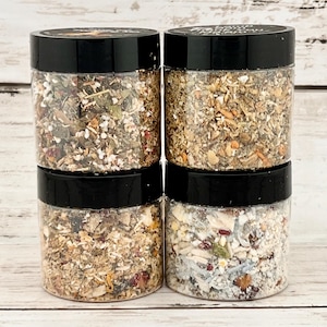 May include: Four small glass jars with black lids filled with different types of dried herbs and flowers. The jars are arranged in two rows of two.