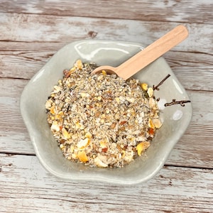 May include: A close-up of a light gray bowl filled with a mixture of dry birdseed. A wooden spoon is resting on top of the birdseed.