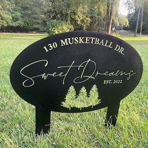 May include: Black metal yard sign with the text "130 MUSKETBALL DR. Sweet Dreams EST. 2022" and a silhouette of three pine trees.