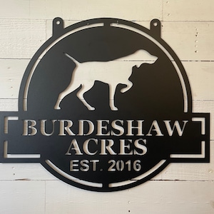 May include: Black metal sign featuring a dog silhouette within a circular frame. The sign reads "BURDESHAW ACRES EST. 2016" in a rectangular panel below the dog. Two small holes are at the top for hanging. The sign is against a white wooden background.