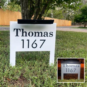May include: A white address sign with the name "Thomas" and the number "1167" in black lettering. The sign has a black light fixture on top and is displayed on a grassy lawn.