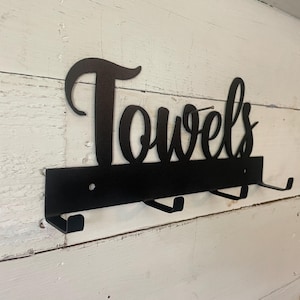 May include: Black metal towel rack with the word "Towels" in a cursive font. The rack has four hooks for hanging towels. The rack is mounted on a white wooden wall.