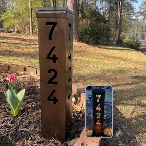May include: A bronze-colored address post with the numbers 7424 cut out. The post is rectangular and stands upright in a garden setting. The numbers are visible on two sides of the post.