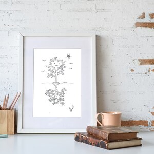 May include: A black and white line drawing of a tree with roots and birds flying above. The drawing is framed in a white frame and is sitting on a white surface with a cup, pencils, and books.