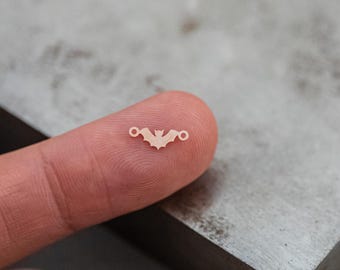 Op de afbeelding: Een kleine, witte metalen vleermuisbedel met twee springringen. De bedel wordt vastgehouden door een vinger.