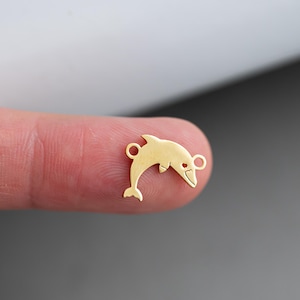 May include: Gold-toned metal dolphin charm with two jump rings. The dolphin is facing right and has a stylized fin.