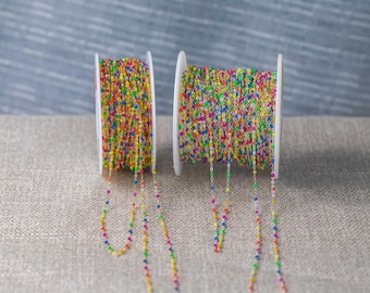 Op de afbeelding: Twee spoelen met kleurrijke kralenketting. De ketting is gemaakt van kleine, ronde kralen in verschillende kleuren, waaronder geel, oranje, roze, blauw en groen. De ketting is geregen op een zilverkleurige draad. De spoelen zijn wit.