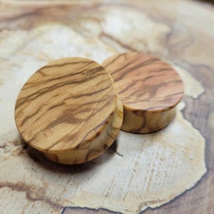 May include: Two round wooden ear gauges with a natural wood grain pattern. The gauges are made of olive wood and have a light brown colour.