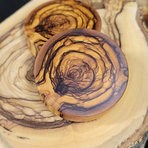 May include: Two round wooden coasters with a natural wood grain pattern. The coasters have a dark brown and light brown colour scheme. The coasters are made of olive wood.