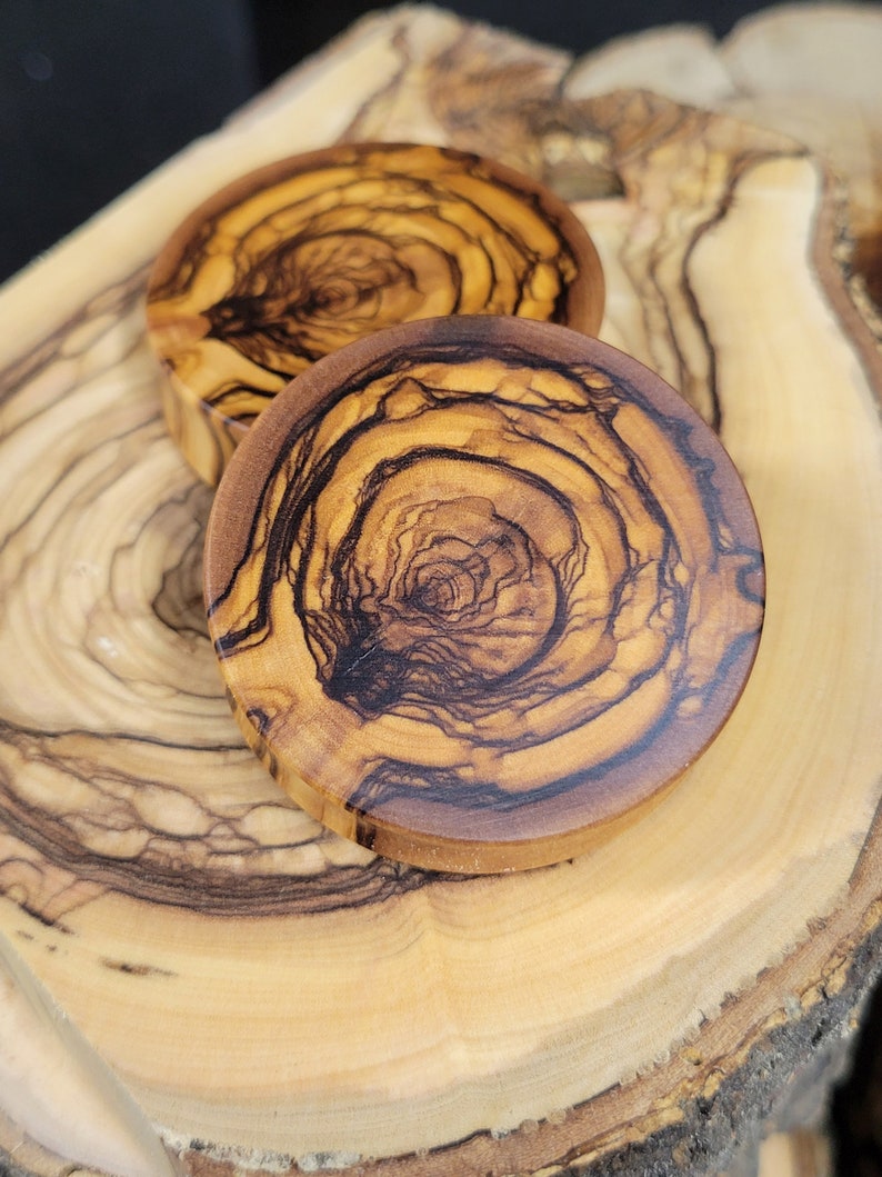 May include: Two round wooden coasters with a natural wood grain pattern. The coasters have a dark brown and light brown color scheme. The coasters are made of olive wood.