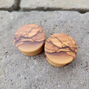 May include: Two round wooden ear gauges with a natural wood grain pattern. The gauges are made of olive wood and have a light brown color.