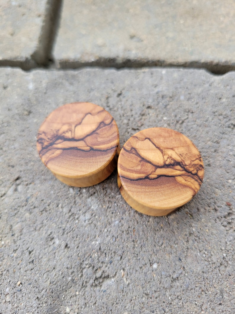 May include: Two round wooden ear gauges with a natural wood grain pattern. The gauges are made of olive wood and have a light brown color.