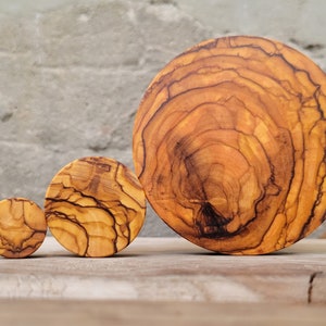 May include: Three round wooden coasters made of olive wood. The coasters are different sizes, with the largest being about 6 inches in diameter. The coasters have a natural wood finish and show the grain of the wood.