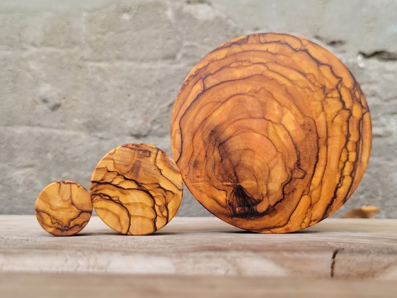 May include: Three round wooden coasters made of olive wood. The coasters are different sizes, with the largest being about 6 inches in diameter. The coasters have a natural wood finish and show the grain of the wood.