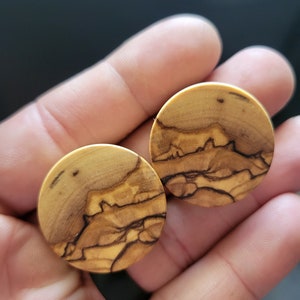 May include: Two round wooden ear gauges with a natural wood grain pattern. The gauges have a light brown color with dark brown streaks.