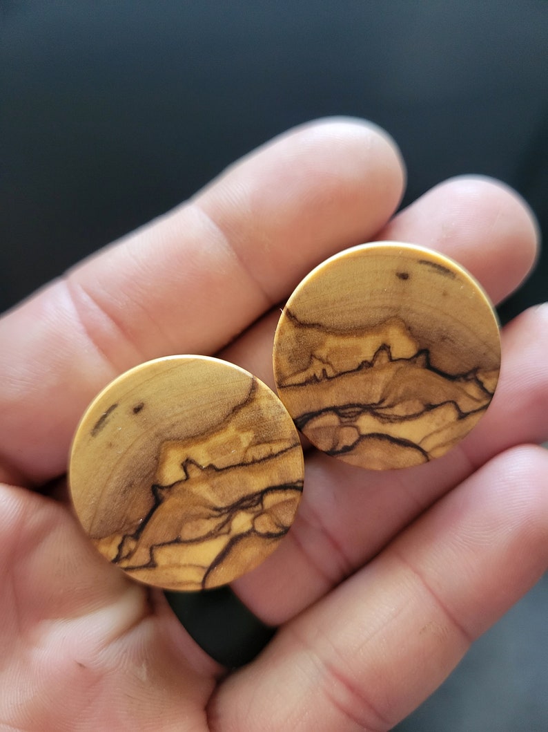 May include: Two round wooden ear gauges with a natural wood grain pattern. The gauges have a light brown color with dark brown streaks.