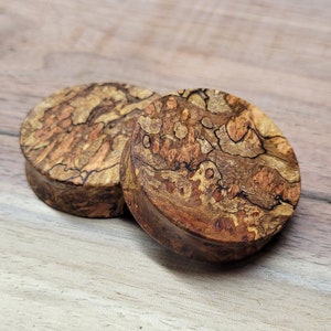 May include: Two round wooden plugs with a unique burl wood grain pattern. The plugs are a mix of brown, tan, and black, with a natural, organic look. The wood has a textured surface and a dark brown edge.