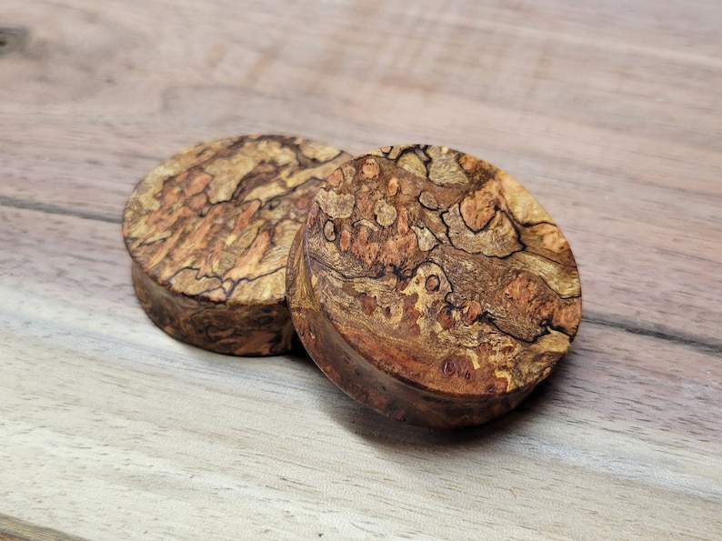 May include: Two round wooden plugs with a unique burl wood grain pattern. The plugs are a mix of brown, tan, and black, with a natural, organic look. The wood has a textured surface and a dark brown edge.