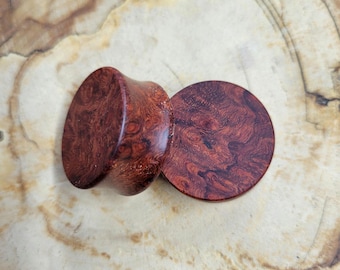 May include: A pair of circular ear plugs crafted from polished wood, featuring a deep reddish-brown colour with a prominent, swirling burl grain pattern. The plugs have a smooth, rounded surface and a cylindrical shape with a flared edge.