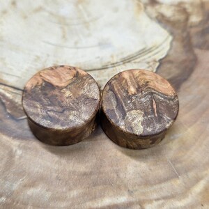 May include: Two wooden ear plugs with a natural wood grain pattern. The plugs are cylindrical with a flat, circular face. The wood features a mix of brown and tan hues, creating a unique, organic appearance. The plugs are displayed on a wooden surface.