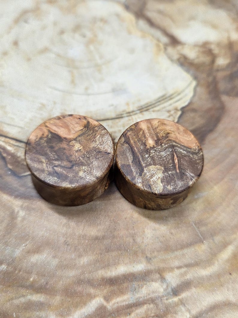 May include: Two wooden ear plugs with a natural wood grain pattern. The plugs are cylindrical with a flat, circular face. The wood features a mix of brown and tan hues, creating a unique, organic appearance. The plugs are displayed on a wooden surface.