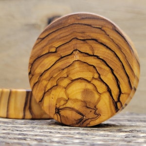 May include: A pair of round wooden ear gauges with a natural wood grain pattern. The gauges have a light brown color with darker brown streaks.