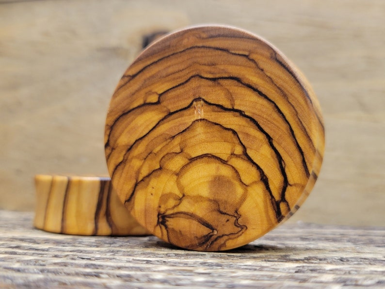 May include: A pair of round wooden ear gauges with a natural wood grain pattern. The gauges have a light brown color with darker brown streaks.