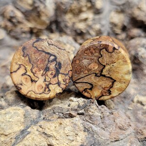 May include: Two wooden ear plugs with a natural, organic pattern. The plugs are round with a light brown base color, and feature dark brown and reddish-brown veining. The wood grain is visible, creating a unique, natural look.