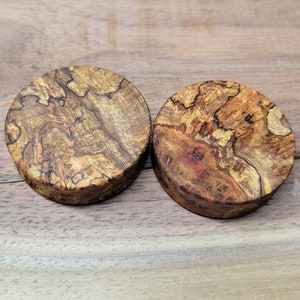 May include: Two round wooden plugs with a natural, textured appearance. The plugs are made of a light brown wood with darker brown and reddish-brown patterns. The wood grain is visible, creating a unique look. The plugs are likely for ear piercing.
