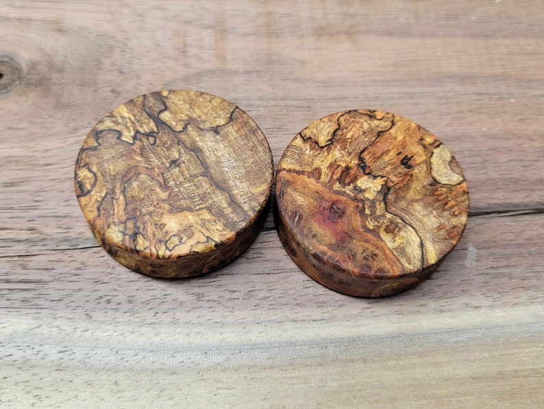 May include: Two round wooden plugs with a natural, textured appearance. The plugs are made of a light brown wood with darker brown and reddish-brown patterns. The wood grain is visible, creating a unique look. The plugs are likely for ear piercing.