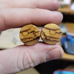 May include: Two round wooden ear gauges with a natural wood grain pattern. The gauges are a light brown color with darker brown streaks.