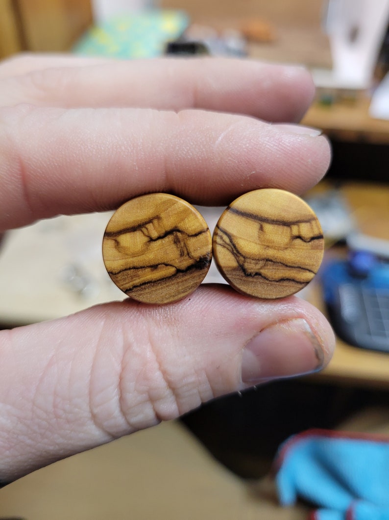 May include: Two round wooden ear gauges with a natural wood grain pattern. The gauges are a light brown color with darker brown streaks.