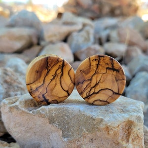 May include: Two round wooden ear gauges with a natural wood grain pattern. The gauges have a dark brown and light brown color scheme.