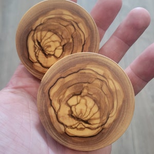 May include: Two round wooden ear gauges with a natural wood grain pattern. The gauges have a light brown color with darker brown streaks.