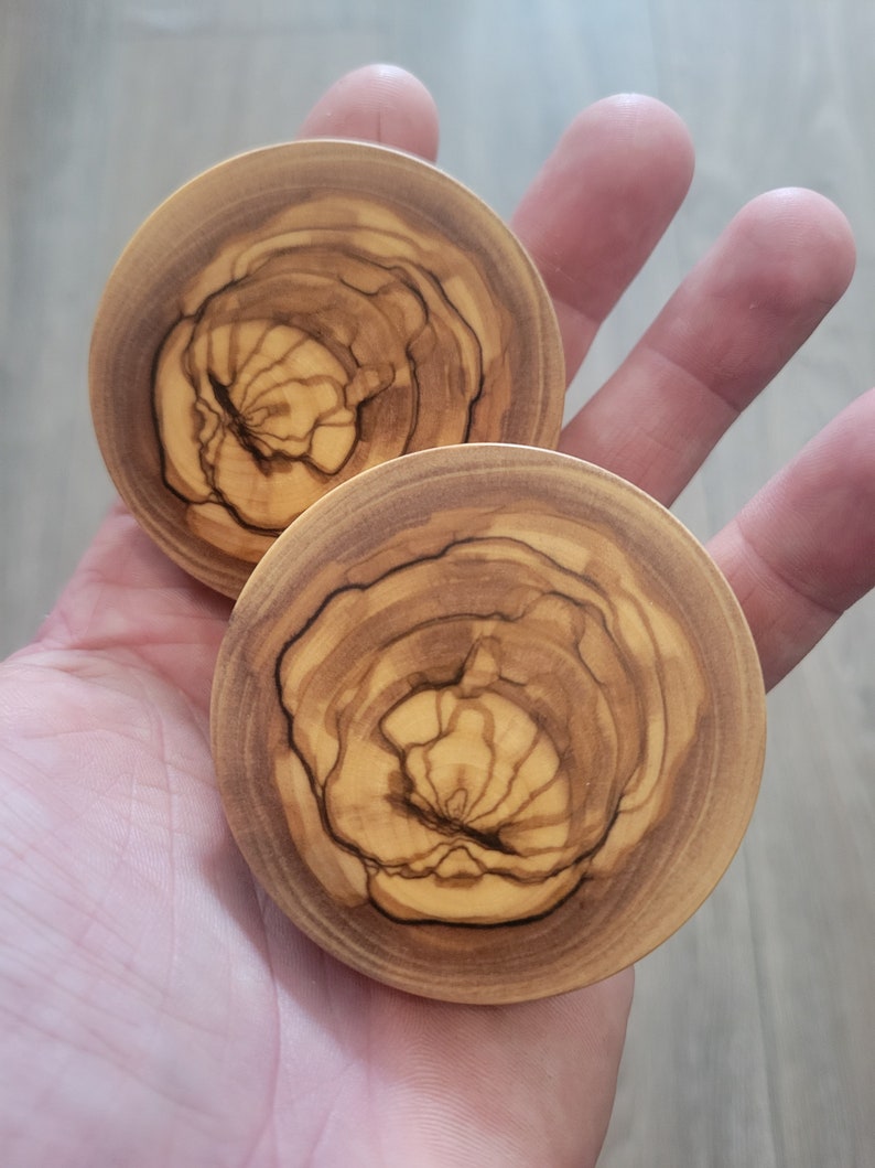May include: Two round wooden ear gauges with a natural wood grain pattern. The gauges have a light brown color with darker brown streaks.