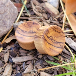 May include: Two round wooden ear gauges with a natural wood grain pattern. The gauges are made of olive wood and have a light brown color.