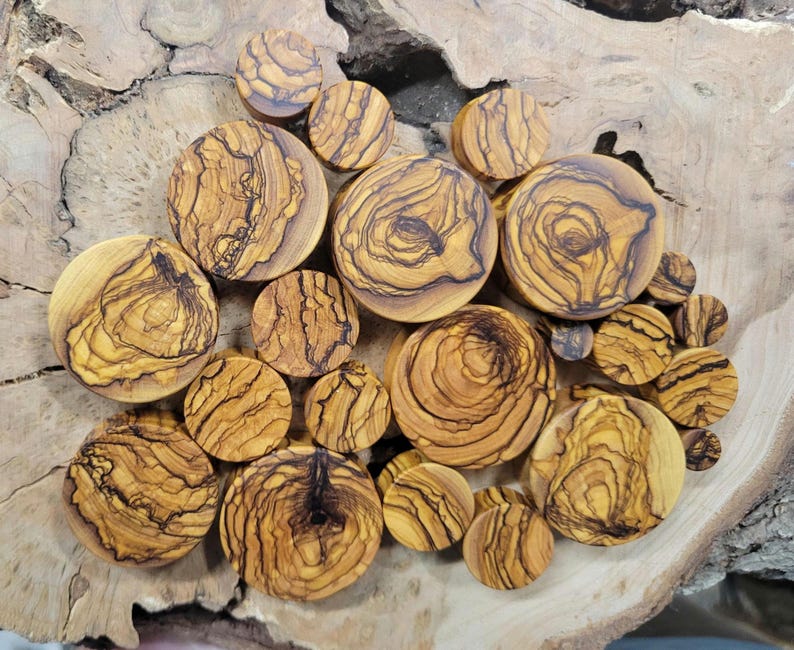 May include: A collection of olive wood ear gauges in various sizes and patterns. The gauges are a natural wood colour with dark brown streaks and swirls.