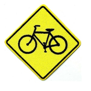 May include: Yellow, diamond-shaped patch featuring a black bicycle symbol. The patch has a black border and is designed to be ironed or sewn onto clothing or accessories. The bicycle graphic is a simple, stylized representation.