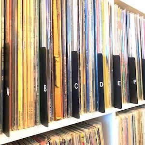以下が含まれることがあります： 白い棚に収納されたレコードのコレクション。AからHまでの黒い仕切りがレコードを区切っています。アルバムのカバーは色とりどりで、テキストやグラフィックが施されたものもあります。レコードは整然と並べられています。