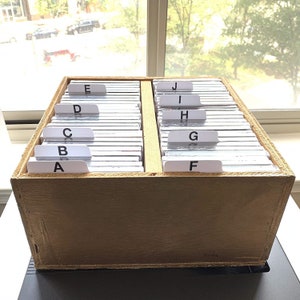 May include: A wooden card catalog box with labeled dividers. The box is divided into two sections, each with multiple cards. The labels are white with black lettering, indicating alphabetical organization. The box is made of light brown wood.